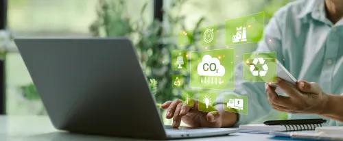 Hände bedienen einen Laptop, vor dem schwebende grüne Symbole zu Nachhaltigkeit, Recycling, Energie und CO₂‑Reduktion angezeigt werden.